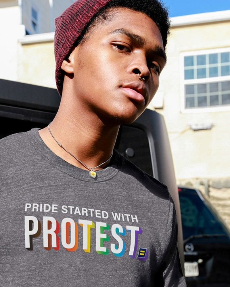 A young Black man in a "Pride Started With Protest" HRC t-shirt stares into the camera.